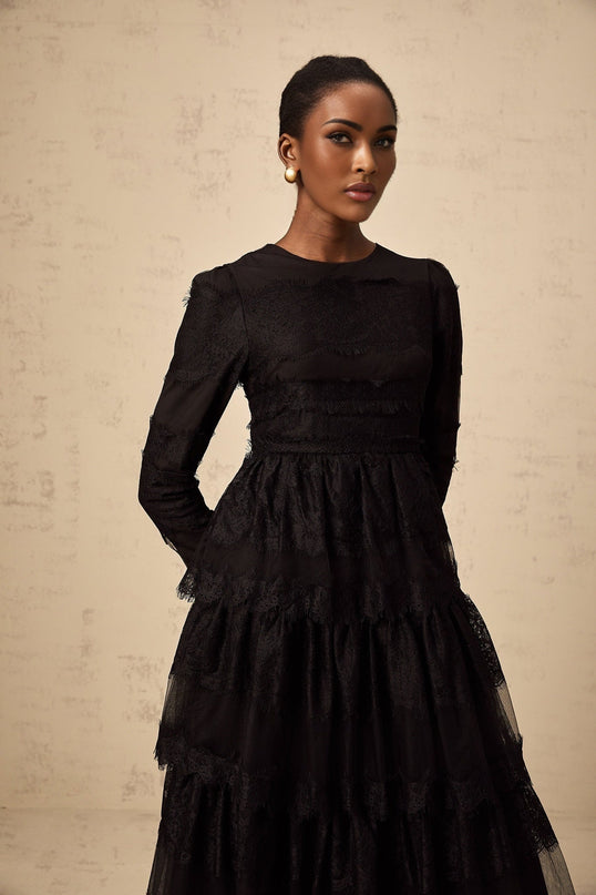 Woman wearing long black lace dress with tiered skirt and sheer tulle details standing against plain background