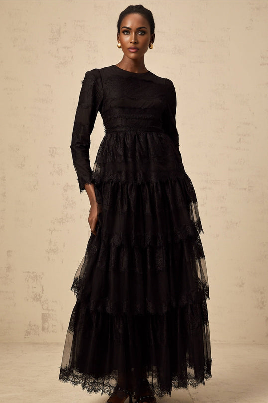 Woman wearing long black lace dress with tiered skirt and sheer tulle details standing against plain background