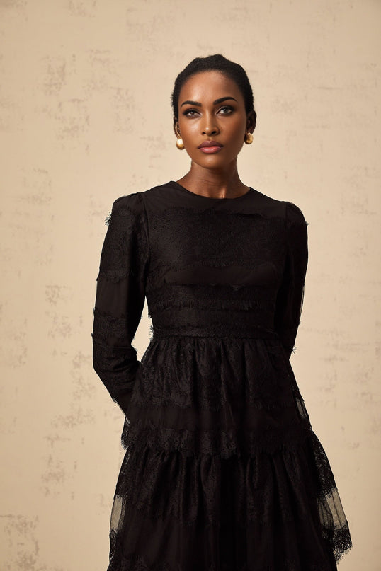 Woman wearing long black lace dress with tiered skirt and sheer tulle details standing against plain background