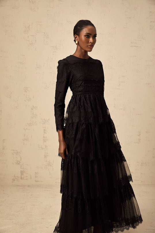 Woman wearing long black lace dress with tiered skirt and sheer tulle details standing against plain background