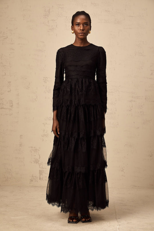 Woman wearing long black lace maxi dress with tiered skirt and sheer tulle details standing against plain background