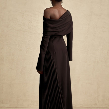 Woman wearing long black pleated maxi dress with high slit posing against plain background