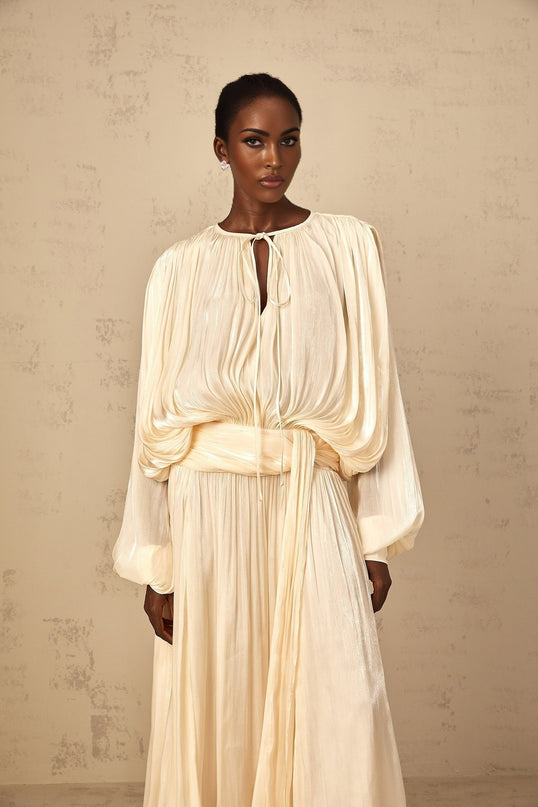 Woman wearing long flowy neutral colored maxi dress with pleated ruffles and sleeveless design standing in outdoor setting