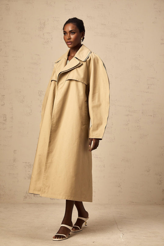 Woman wearing long khaki trench coat with curved sleeves and belted waist standing against plain background
