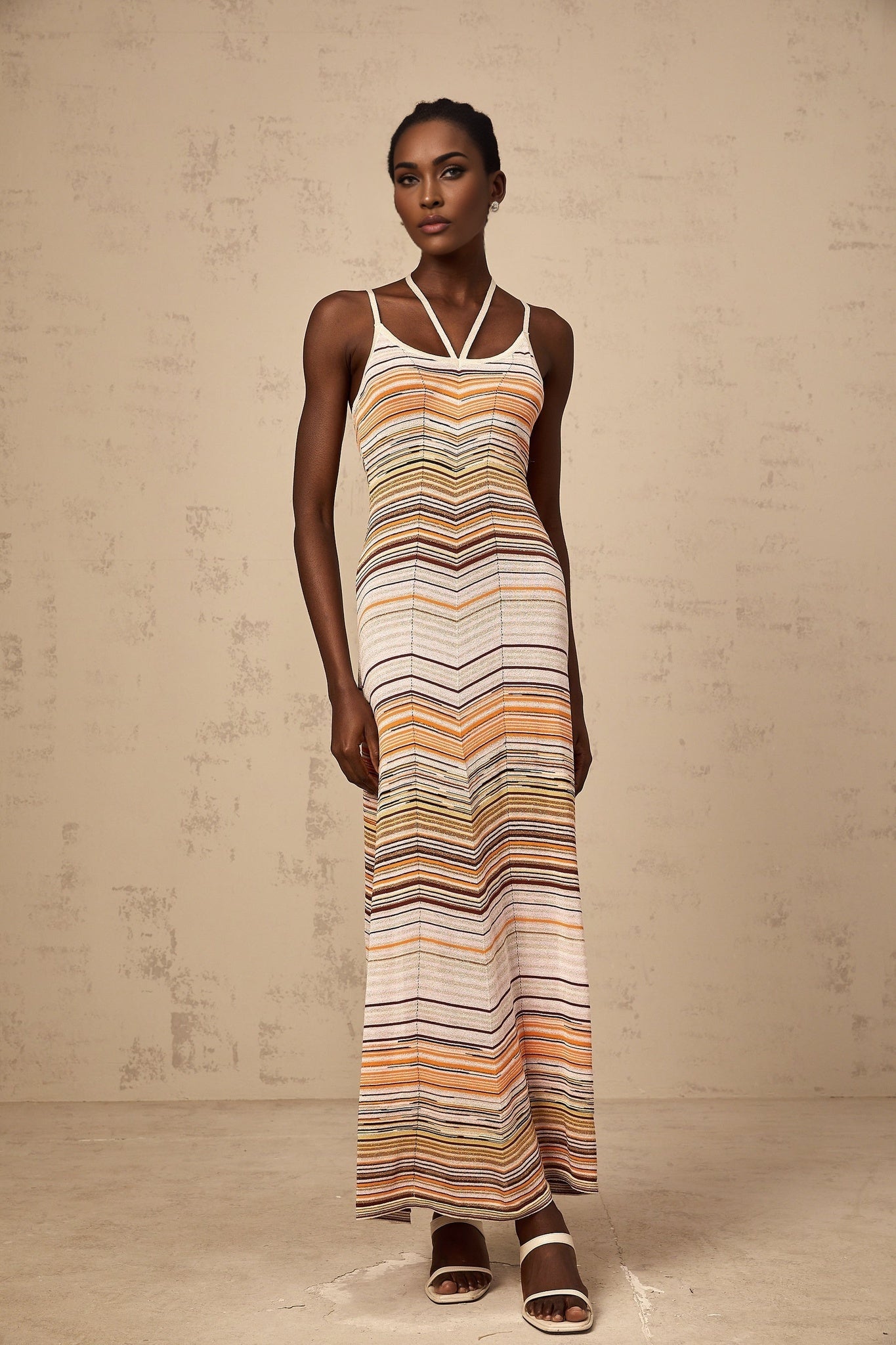 Woman wearing long striped knit maxi dress with multicolored horizontal stripes in pastel shades standing against neutral background