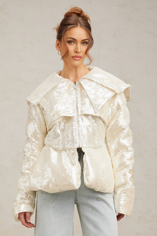 Woman wearing long white down coat with sequin details standing against plain background