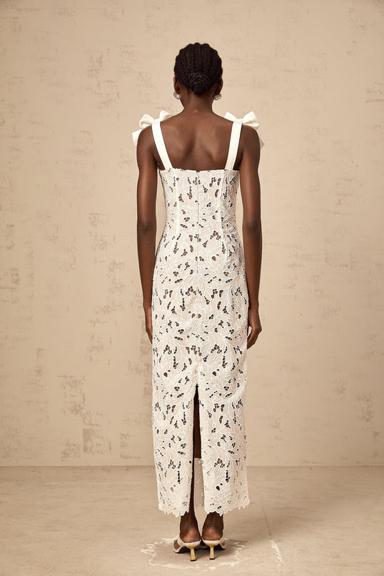 Woman wearing long white maxi dress with floral embroidery detail standing against plain background