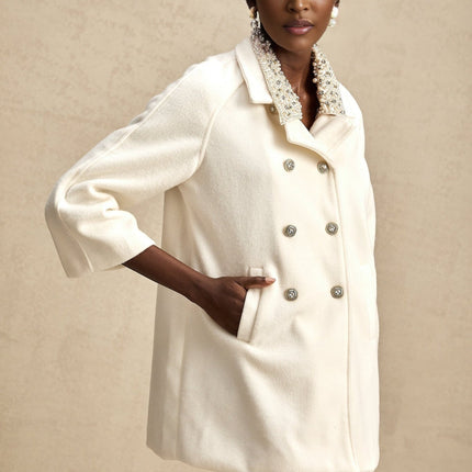 Woman wearing long white woolen coat with embellished details standing against neutral background