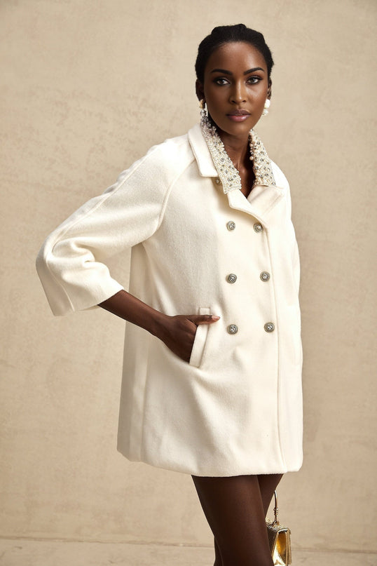 Woman wearing long white woolen coat with embellished details standing against neutral background