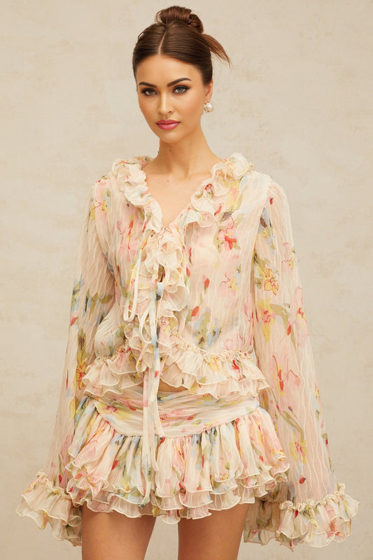 Woman wearing matching floral print blouse and skirt set Blouse has ruffled details Skirt falls below knees Both pieces feature colorful floral pattern on light background