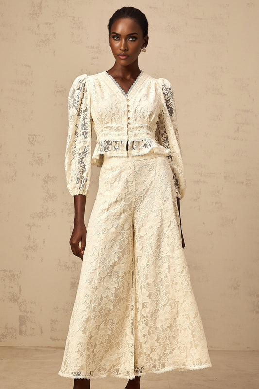 Woman wearing matching neutral colored floral lace blouse and trousers set standing against plain background