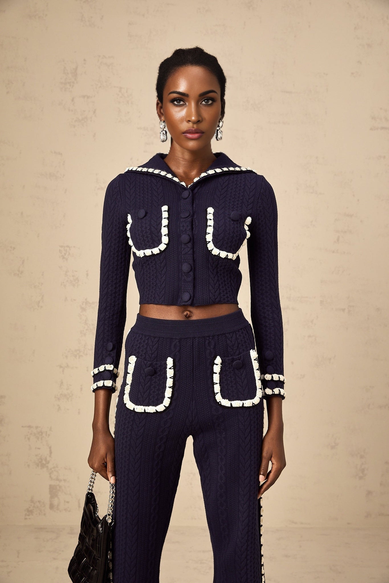 Woman wearing navy blue crochet knit cardigan and matching trousers set standing against plain background
