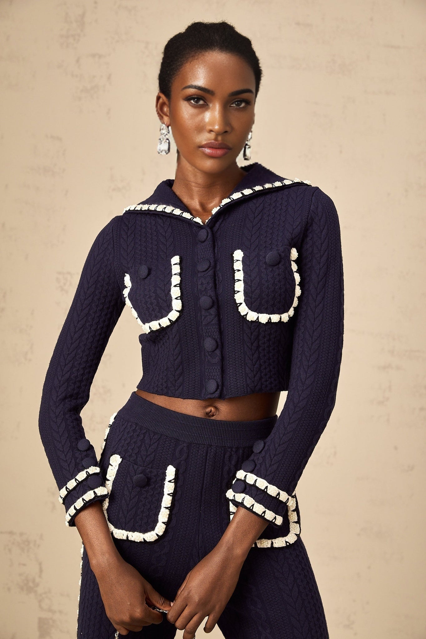 Woman wearing navy blue crochet knit cardigan and matching trousers standing against plain background