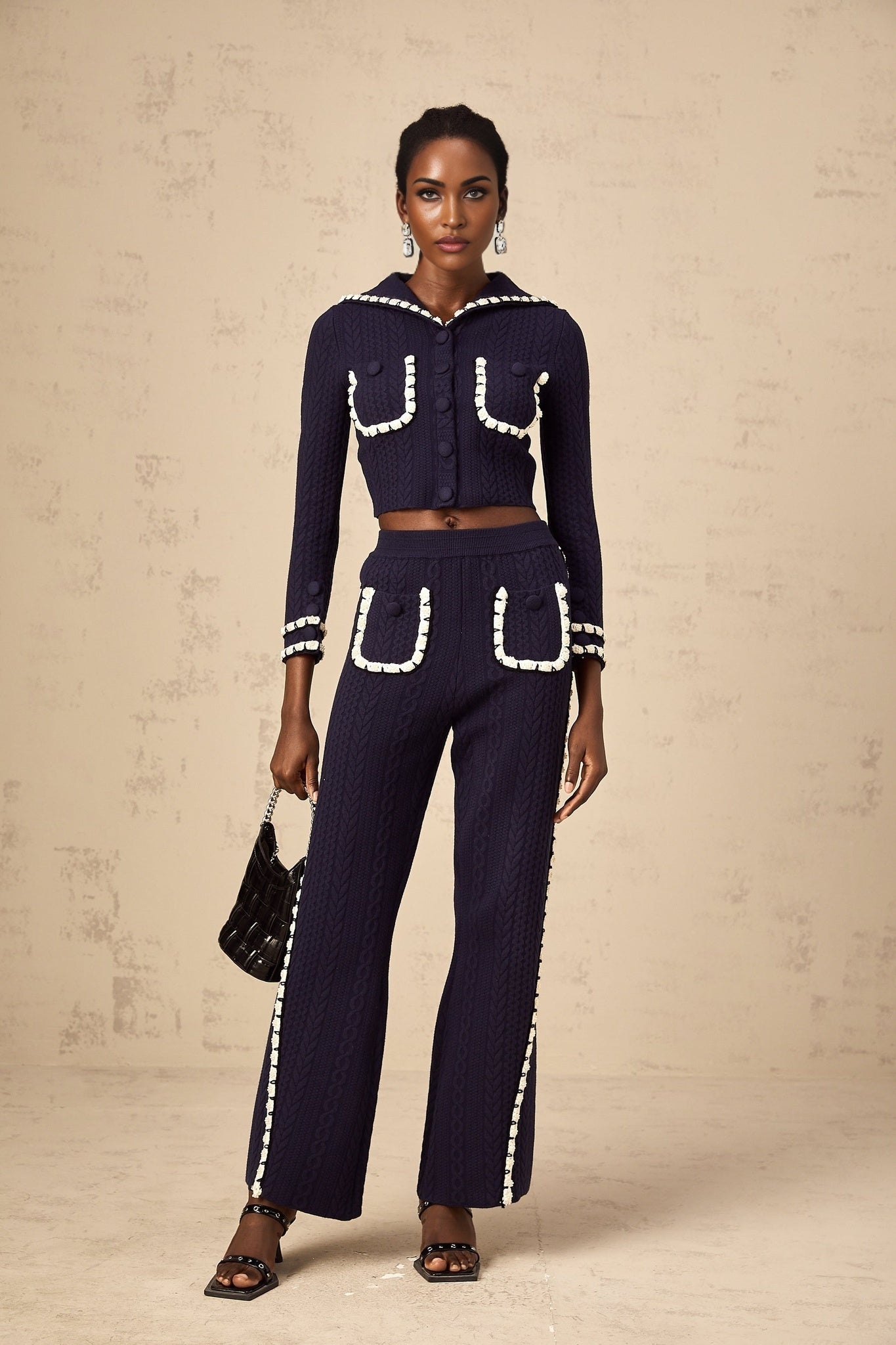 Woman wearing navy blue crochet knit cardigan and matching trousers standing against plain background