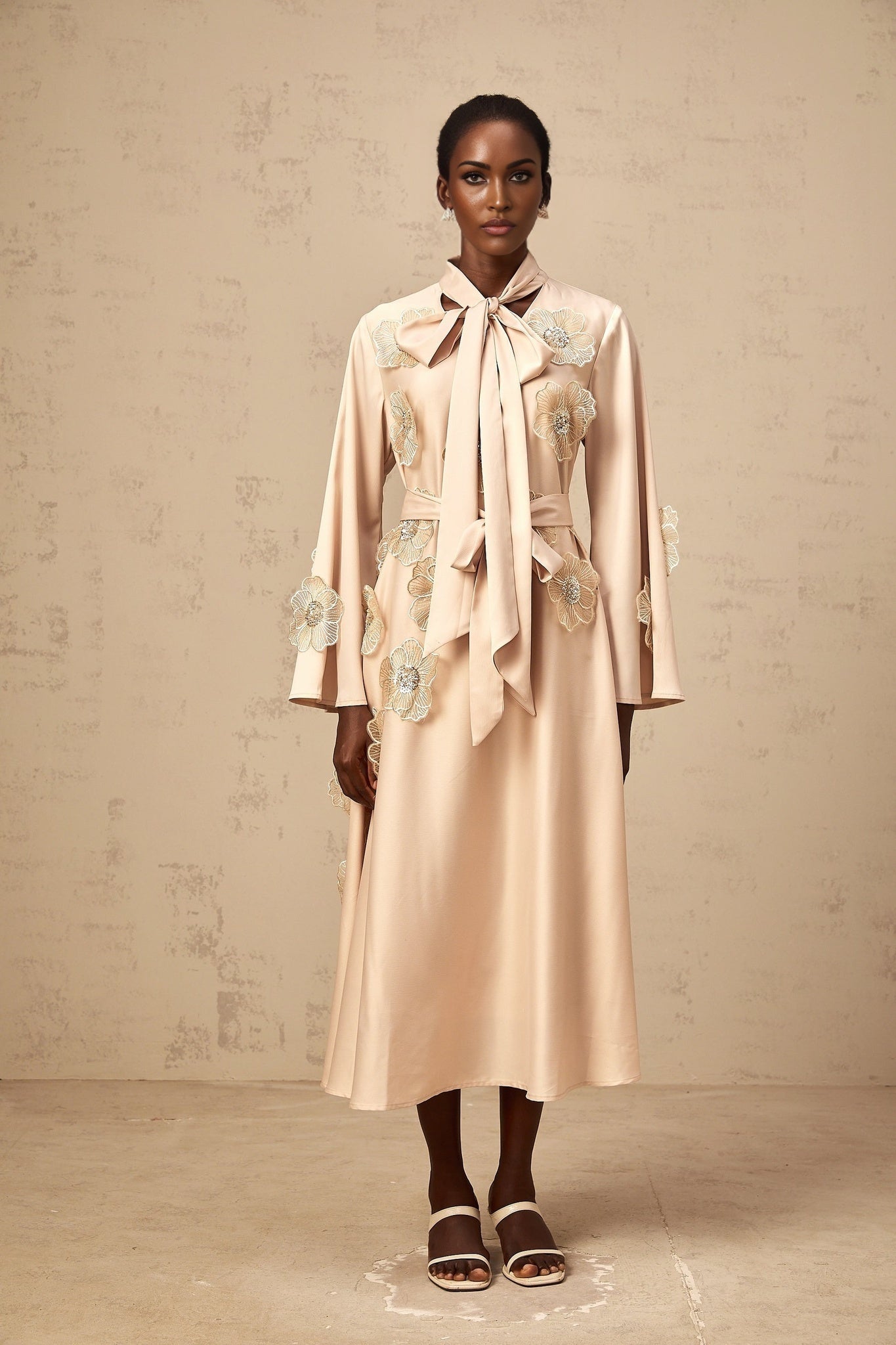 Woman wearing neutral colored floral midi dress with tie detail and flared skirt floral applique pattern visible on dress fabric