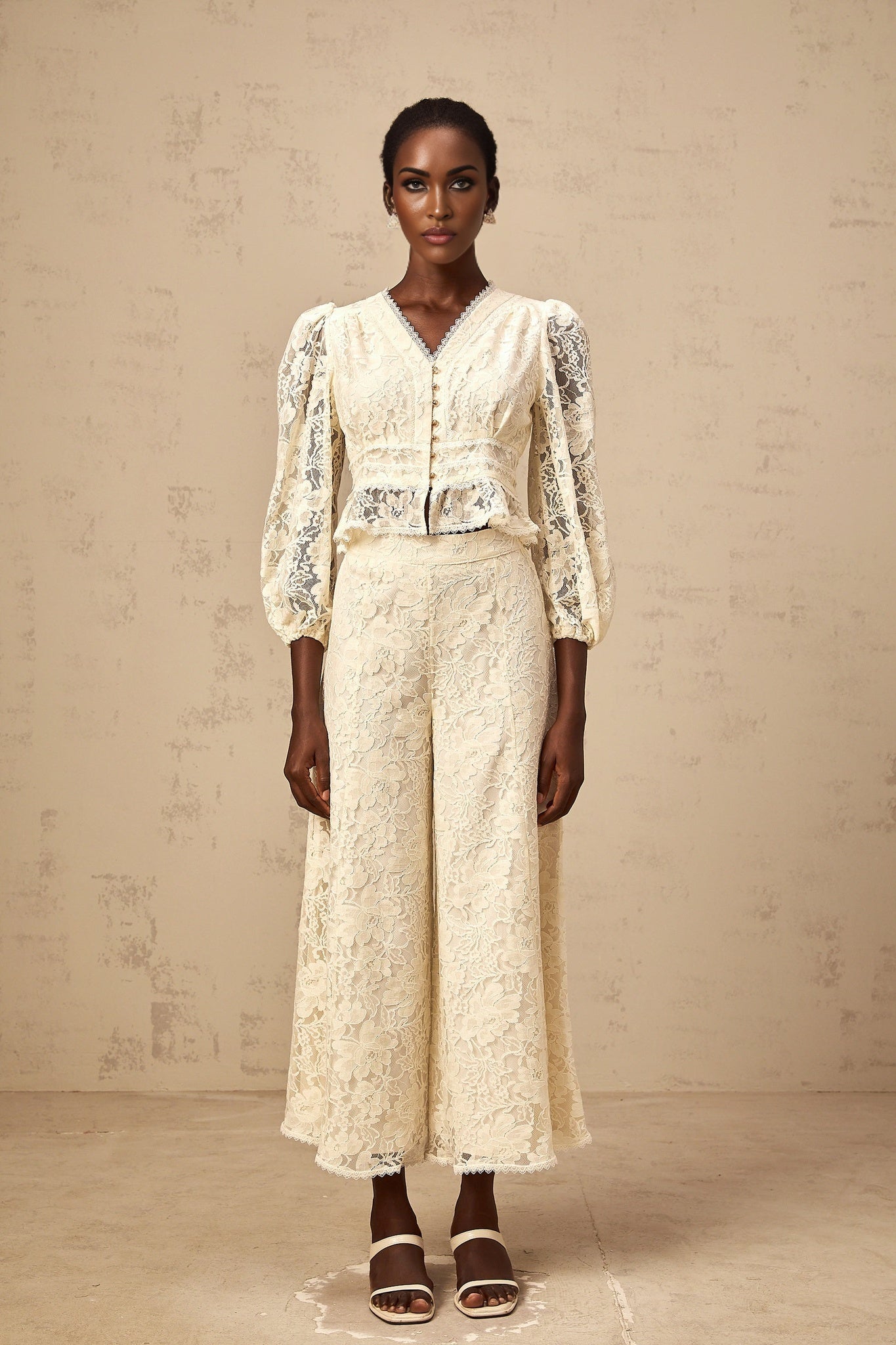 Woman wearing neutral colored matching set with floral lace blouse and trousers standing in relaxed pose