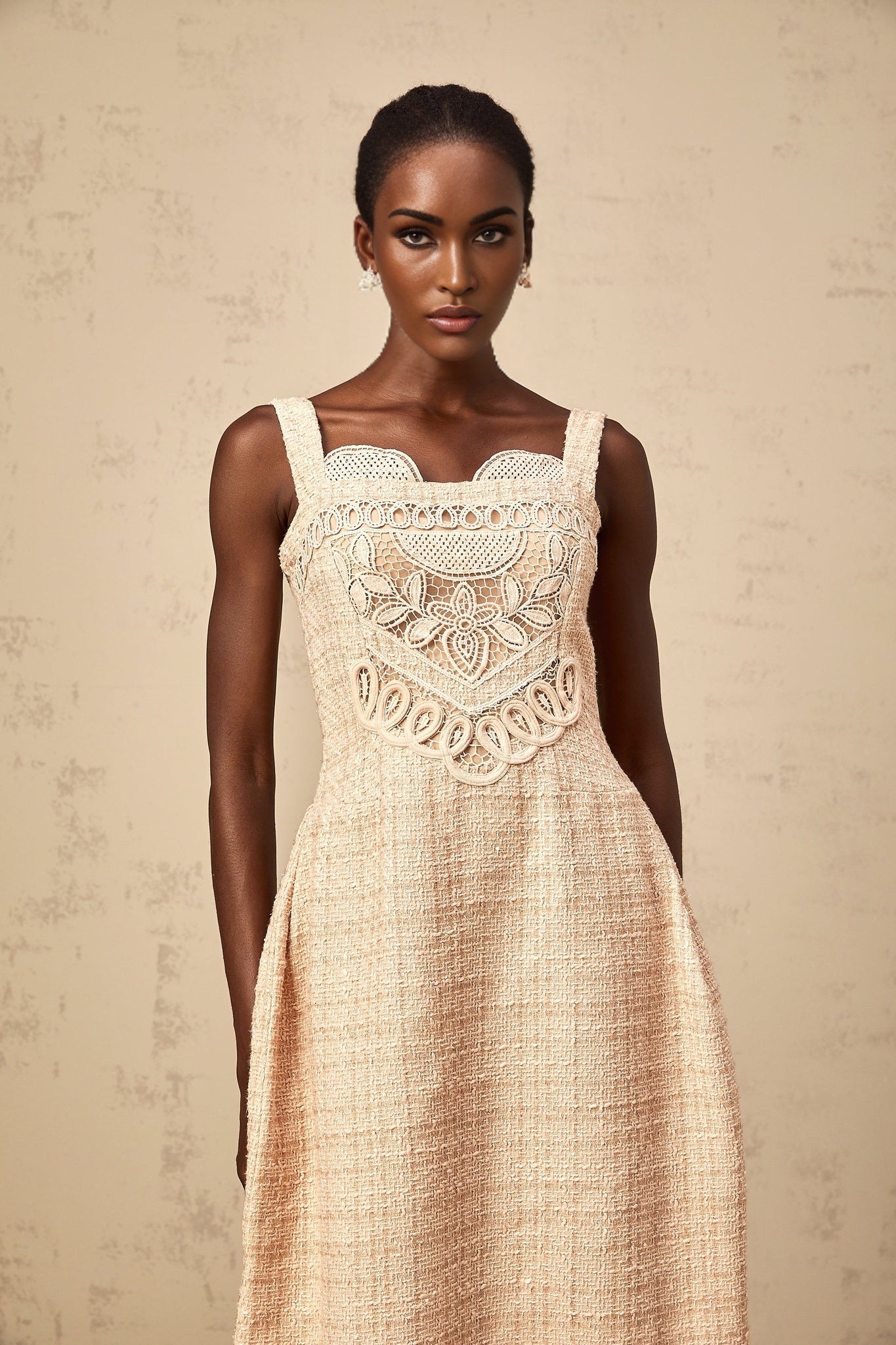 Woman wearing short beige dress with intricate embroidery pattern standing against plain background