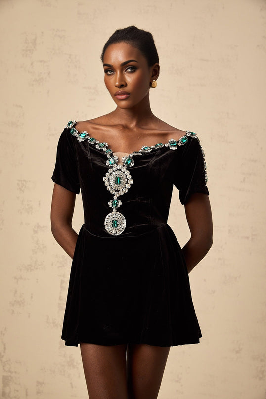 Woman wearing short black velvet dress with embellishments and thin straps standing against plain background
