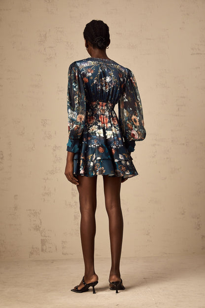 Woman wearing short blue dress with graphic print pattern and embellishments standing in relaxed pose against plain background