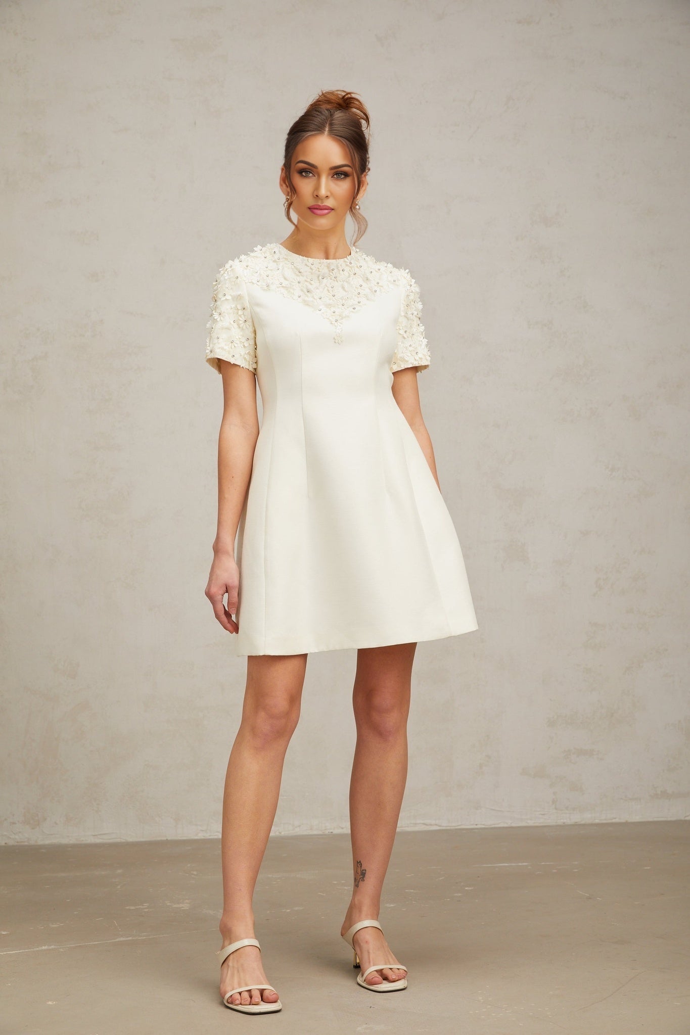 Woman wearing short white dress with colorful floral appliques standing against plain background