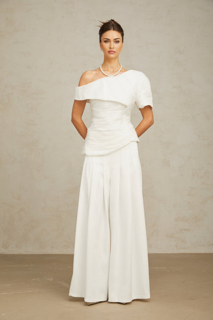 Woman wearing white creased top and matching trousers standing against plain background