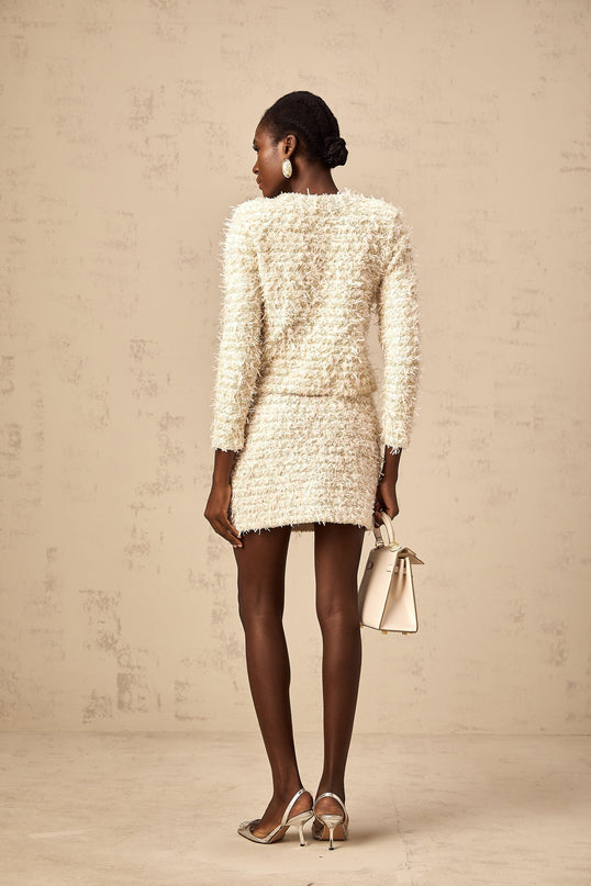 Woman wearing white fuzzy knit cardigan and matching skirt set standing against plain background