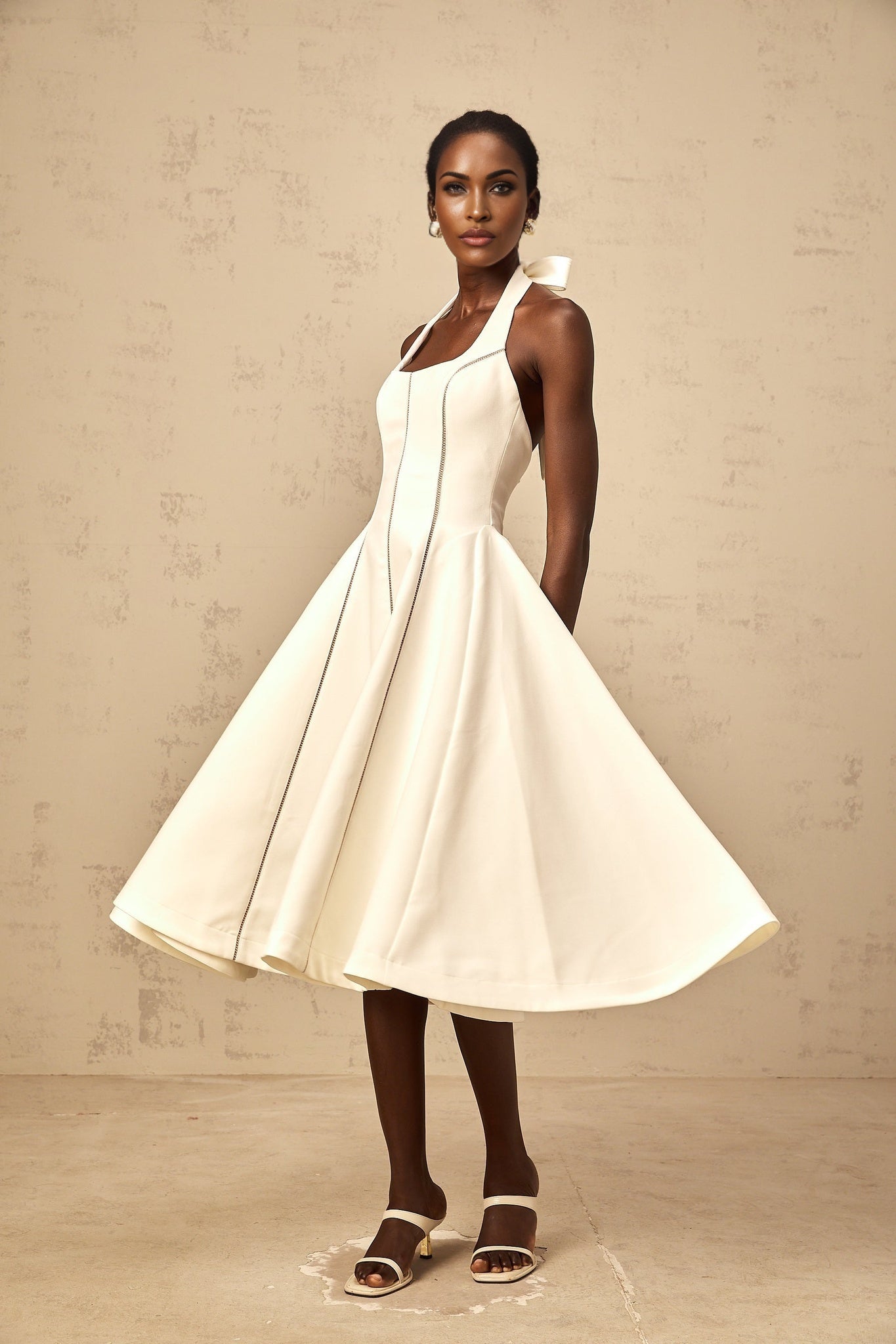 Woman wearing white halterneck midi dress with bow detail at neck standing against plain background