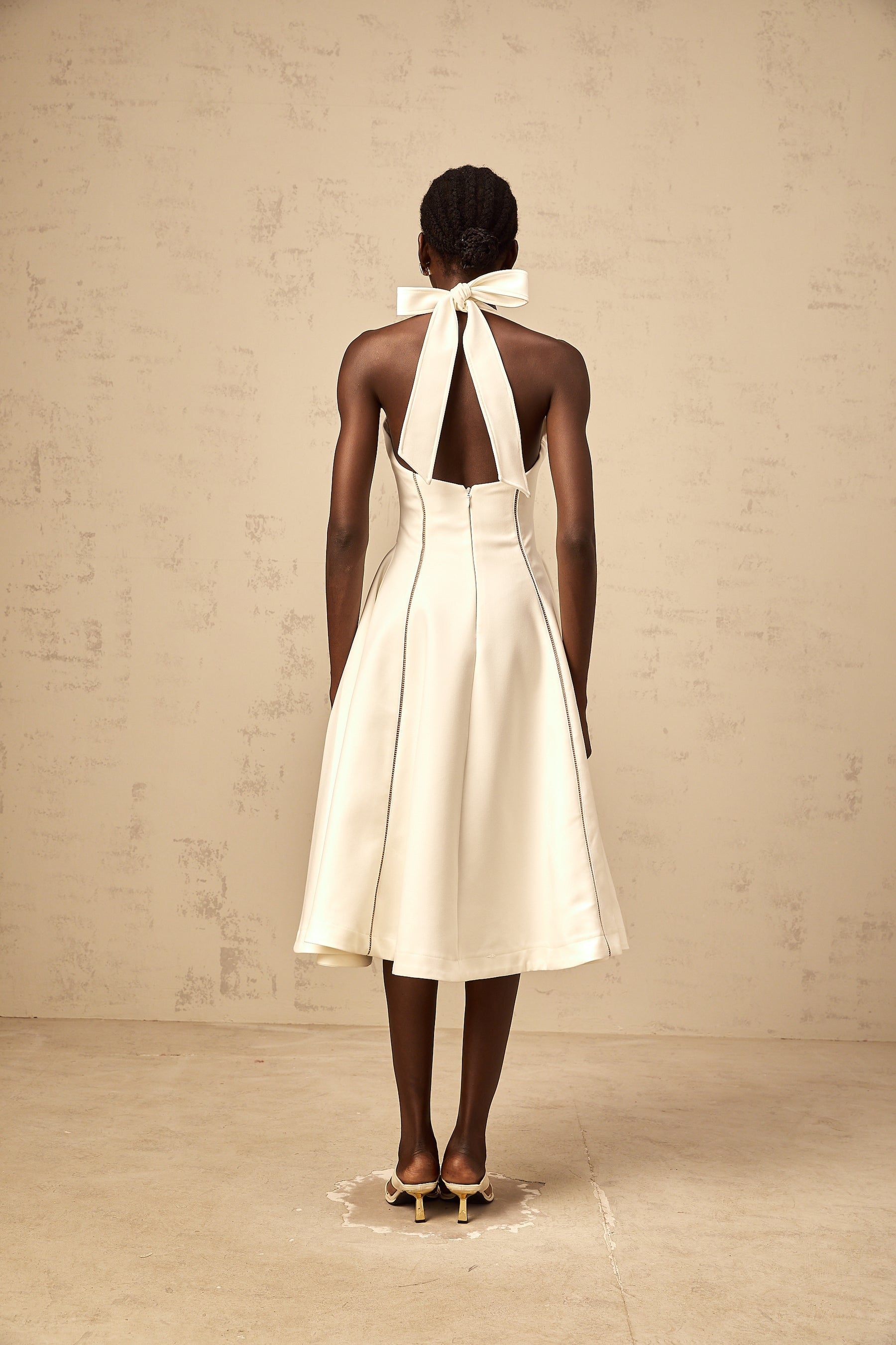 Woman wearing white halterneck midi dress with bow detail at neckline standing against plain background