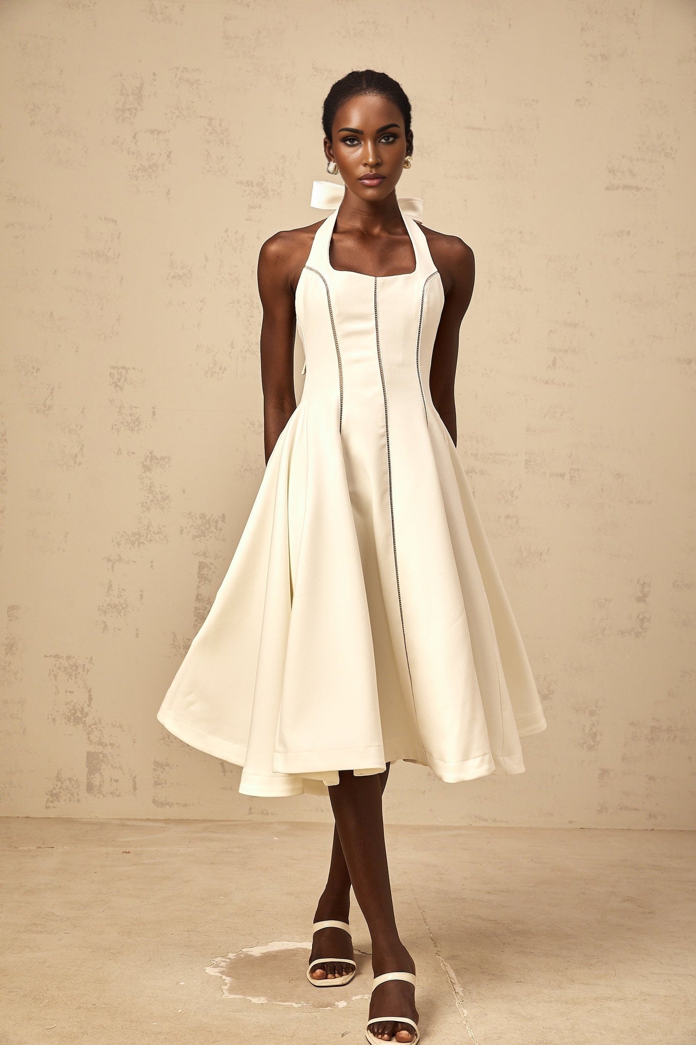Woman wearing white halterneck midi dress with bow detail at the neck standing against plain background