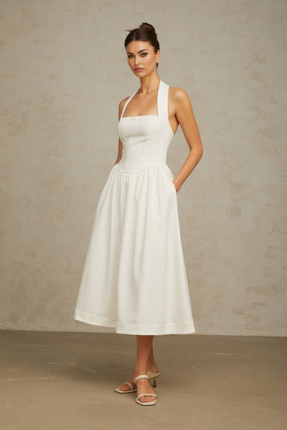 Woman wearing white halterneck midi dress with pleated skirt and fitted bodice standing against plain background