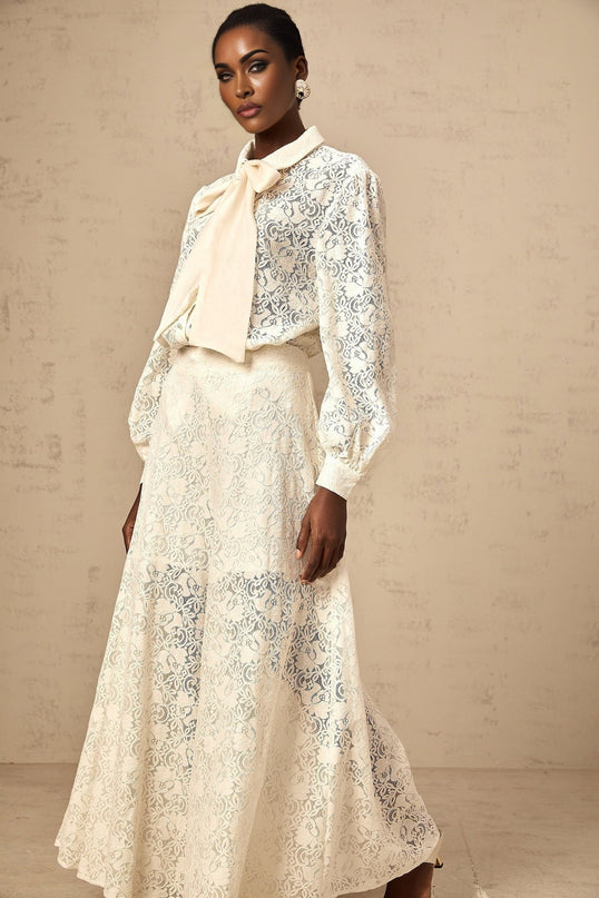 Woman wearing white lace shirt and matching trousers set against plain background