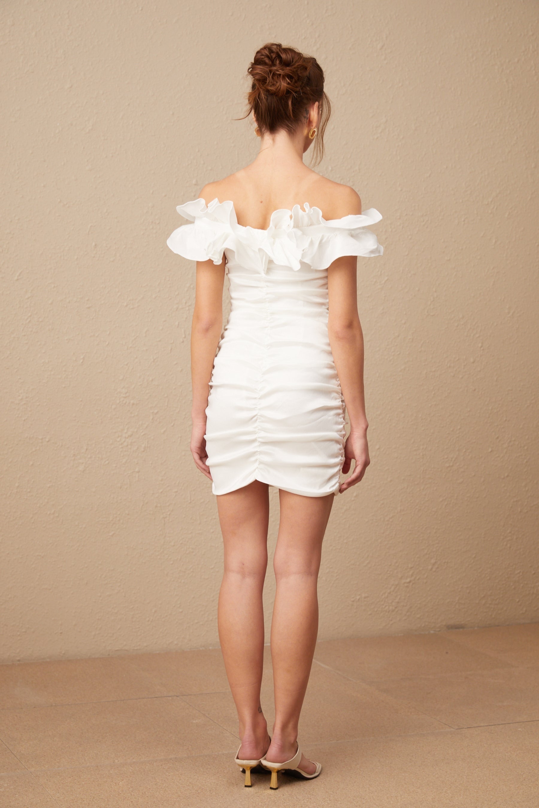 Woman wearing white off shoulder mini dress with ruffled details standing against plain background