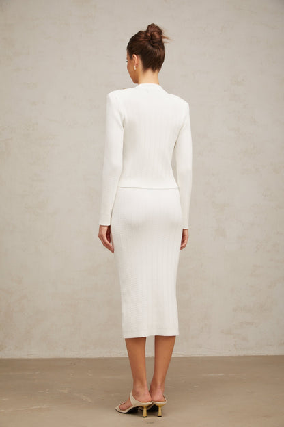 Woman wearing white ribbed knit jacket and matching skirt set standing against plain background