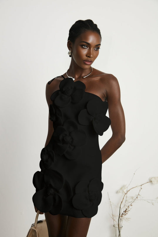 Young woman wearing a black floral offshoulder mini dress with a liane pattern standing against a plain background