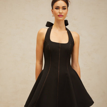 Young woman wearing a black halterneck mini dress with bow detail at the neck standing against a plain background