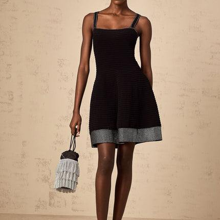 Young woman wearing a black knit mini dress with flared skirt and studded details standing in front of plain background