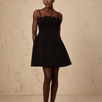 Young woman wearing a black mini dress with flared skirt and rose applique details standing against plain background