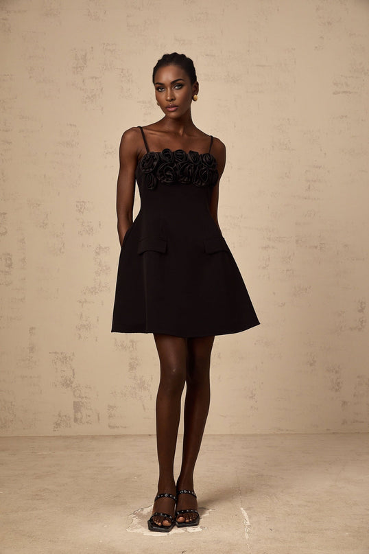 Young woman wearing a black mini dress with flared skirt and rose applique details standing against plain background