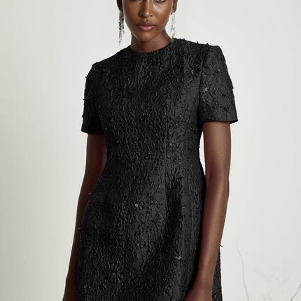 Young woman wearing a black mini dress with floral applique embellishments standing against a plain background