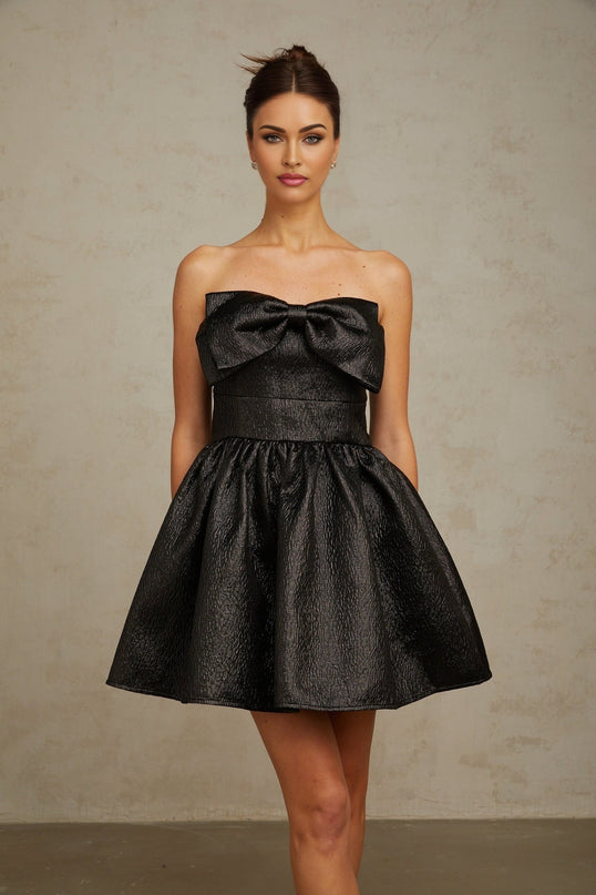 Young woman wearing a black mini dress with metallic accents and a large bow detail standing against a plain background