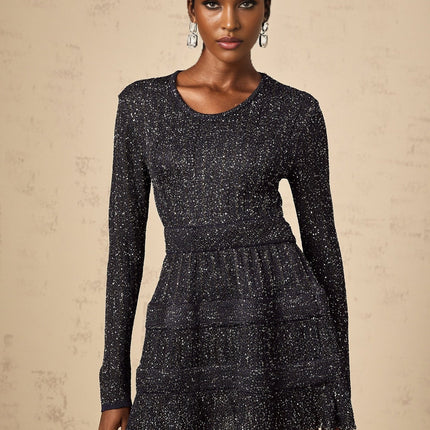 Young woman wearing a black sequined knitted mini dress with long sleeves standing against a plain background