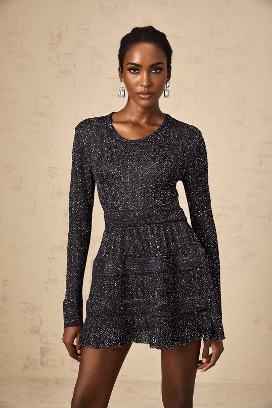 Young woman wearing a black sequined knitted mini dress with long sleeves standing against a plain background