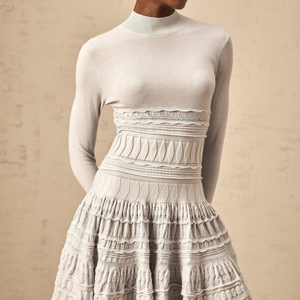 Young woman wearing a grey mini dress with ruffled details and ribbed texture standing against a plain background