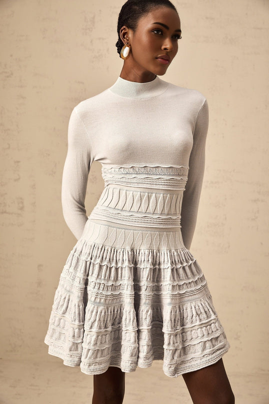 Young woman wearing a grey mini dress with ruffled details and ribbed texture standing against a plain background