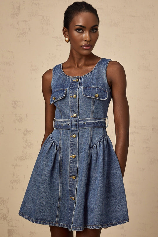 Young woman wearing a light blue denim mini dress with a belted waist and short sleeves standing against a plain background