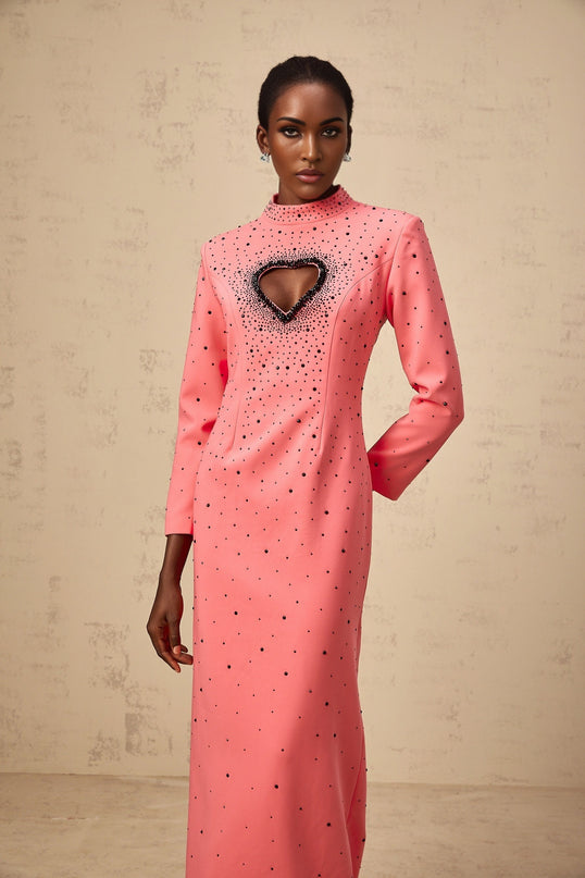 Young woman wearing a long pink maxi dress with heart shaped cutouts and embellishments standing in a neutral background