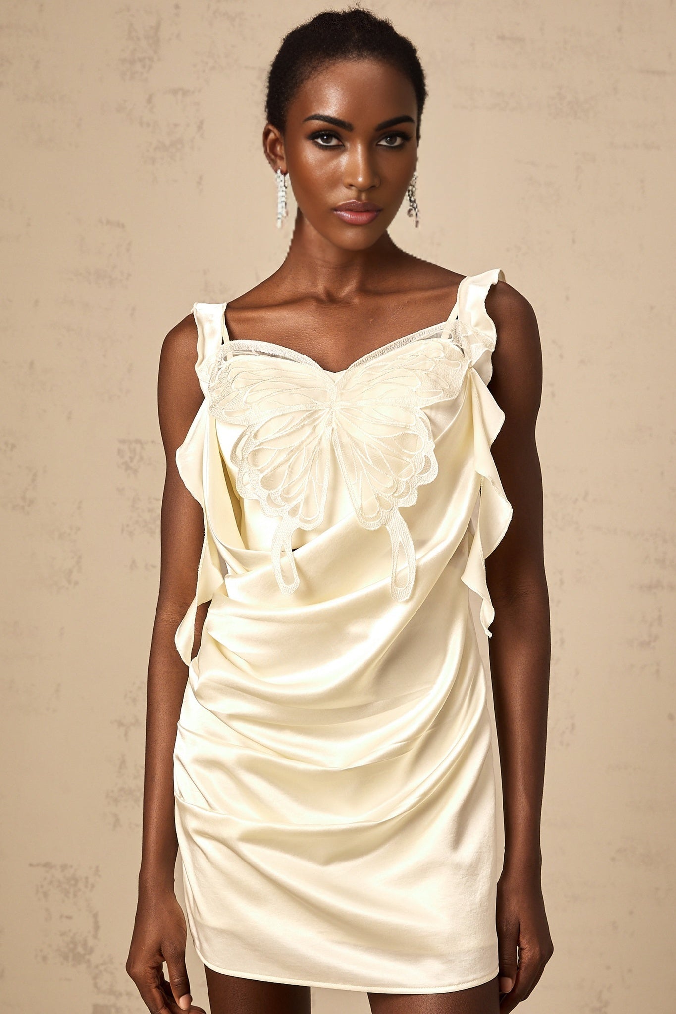 Young woman wearing a neutral colored mini dress with butterfly print and draped design standing in front of plain background