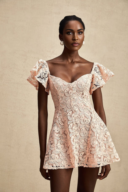 Young woman wearing a pink lace mini dress with flutter sleeves standing against a plain background