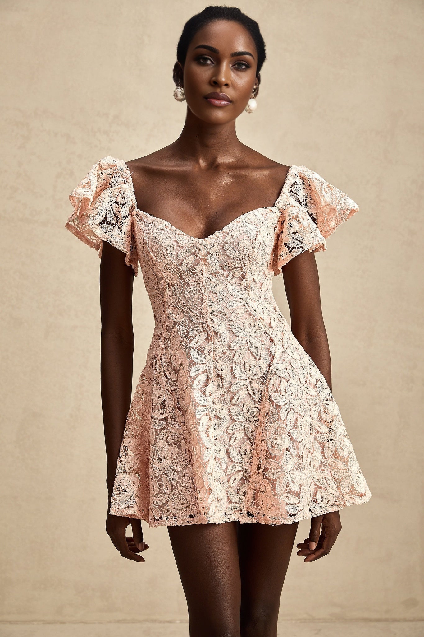 Young woman wearing a pink lace mini dress with flutter sleeves standing in front of a plain background