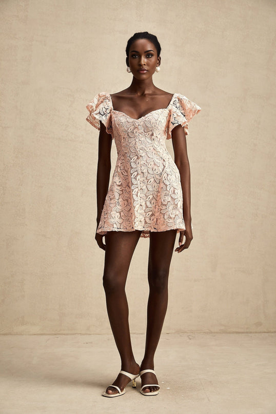 Young woman wearing a pink lace mini dress with flutter sleeves standing in front of a plain background